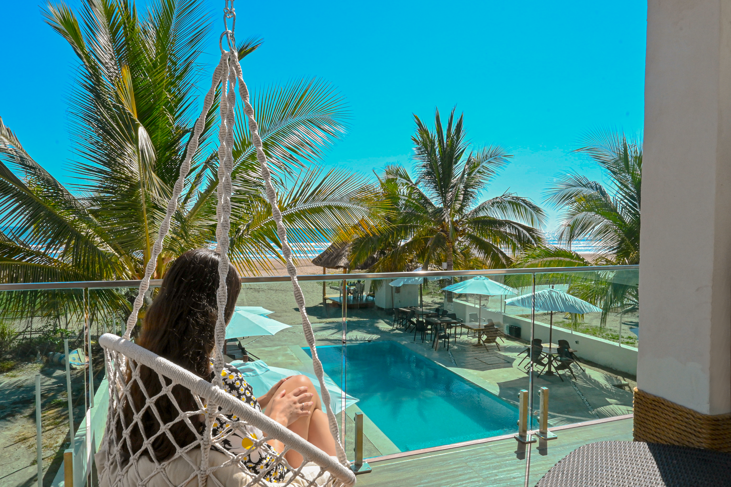 Vista al mar desde el balcón Modelo sentada en columpio colgante disfrutando la vista a la alberca, palmeras y playa desde un balcón.