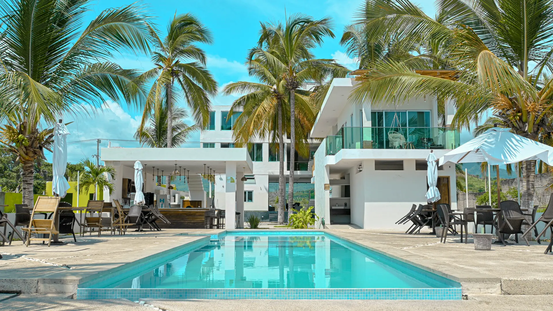 Vista frontal del hotel con piscina, palmeras, área de restaurante y balcones, bajo un cielo azul despejado.